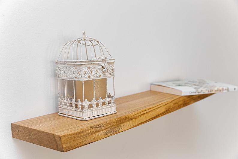 May include: A white birdcage with intricate details sits on a wooden shelf with a book on top. The shelf is mounted on a white wall.