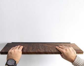 Puede incluir: Una persona instalando una estantería de madera marrón oscuro con soportes de metal negro en una pared blanca.