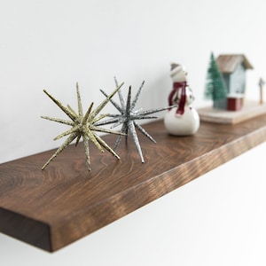 Pode incluir: Uma prateleira de madeira com uma figura de boneco de neve, uma pequena &aacute;rvore de folha perene e uma figura de casa. H&aacute; tamb&eacute;m dois enfeites em forma de estrela brilhantes, um dourado e outro prateado.