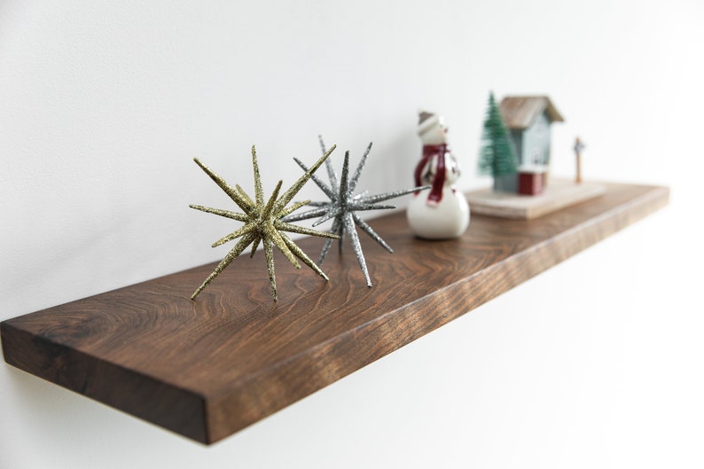 Pode incluir: Uma prateleira de madeira com uma figura de boneco de neve, uma pequena &aacute;rvore de folha perene e uma figura de casa. H&aacute; tamb&eacute;m dois enfeites em forma de estrela brilhantes, um dourado e outro prateado.