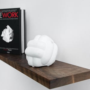 Pode incluir: Uma escultura de cer&acirc;mica branca em forma de n&oacute; est&aacute; sobre uma prateleira de madeira marrom escura. Um livro preto intitulado "REWORK" est&aacute; na prateleira atr&aacute;s da escultura.