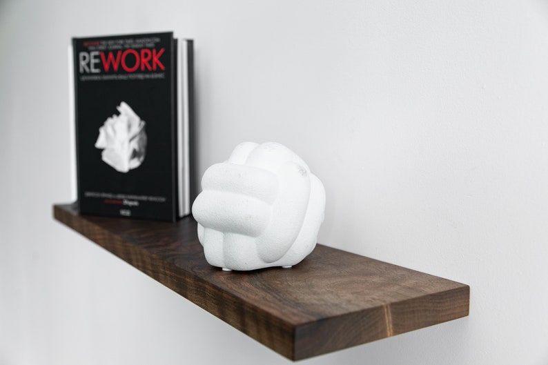 Pode incluir: Uma escultura de cer&acirc;mica branca em forma de n&oacute; est&aacute; sobre uma prateleira de madeira marrom escura. Um livro preto intitulado "REWORK" est&aacute; na prateleira atr&aacute;s da escultura.