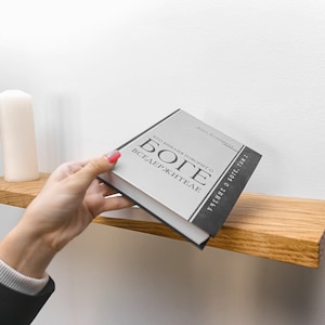 May include: A hand reaches for a gray book titled "ЧТО МИЛАЯ ЛЮБОВЬ  О  БОГЕ ВСЕДЕРЖИТЕЛЕ" with black text on a wooden shelf. A white candle sits on the shelf.