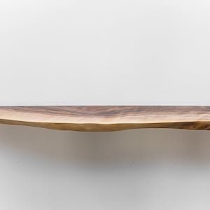 May include: A natural wood shelf with a live edge, the shelf is brown with a light brown wood grain. The shelf is mounted to a white wall.