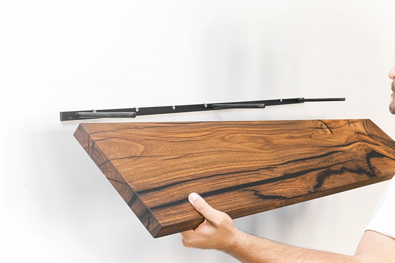 May include: A person installing a wooden shelf with black metal brackets. The shelf is made of dark wood with a natural grain pattern. The brackets are attached to the wall with screws.