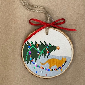 May include: A round wooden Christmas ornament with a green tree and a cat with orange fur. The tree has colorful lights and a star on top. The cat is walking under the lights.