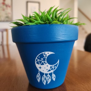 Puede incluir: Una maceta de terracota azul con un diseño de luna creciente y plumas blancas. La maceta está llena de hierba artificial verde.