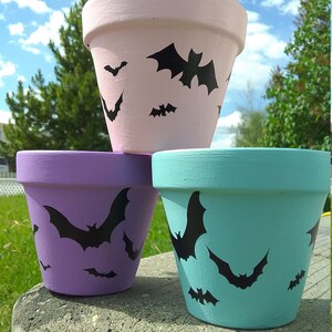 May include: Three pastel-coloured terracotta flower pots, painted pink, purple, and teal. Each pot features black bat silhouettes, creating a Halloween theme. The pots are stacked on a concrete surface.