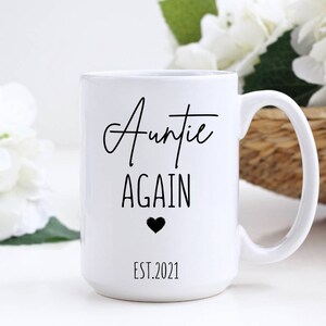 May include: White ceramic mug with the words "Auntie Again" in black script, a black heart, and the year "EST.2021". The mug has a large handle and is surrounded by white flowers.