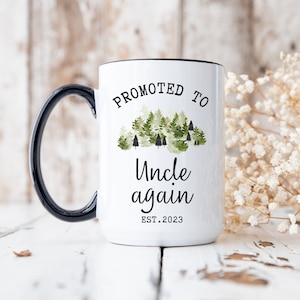May include: White ceramic mug with a black handle. The mug has a black rim and says "Promoted to Uncle again EST. 2023" with a watercolor forest design.