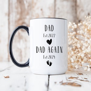 May include: White ceramic coffee mug with a black handle. The mug has black text that reads "Dad Est. 2022" with a small heart and "Dad Again Est. 2024" with a footprint symbol.