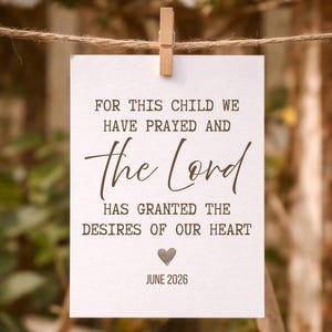 Op de afbeelding: Een witte kaart met de tekst "FOR THIS CHILD WE HAVE PRAYED AND the Lord HAS GRANTED THE DESIRES OF OUR HEART JUNE 2026" is met een houten knijper aan een touwtje geklemd. De achtergrond is wazig met groen en witte bloemen.