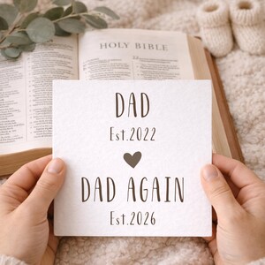 Op de afbeelding: Een witte kaart met de tekst "DAD Est.2022" en "DAD AGAIN Est.2026" met een hartsymbool. De kaart wordt in handen gehouden, met een Heilige Bijbel en babyslofjes op de achtergrond. De scène bevindt zich op een zacht, getextureerd oppervlak.