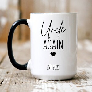 May include: White ceramic mug with a black handle and black rim. The mug has black text that reads "Uncle Again" with a small black heart below the text. The text "EST. 2021" is below the heart.