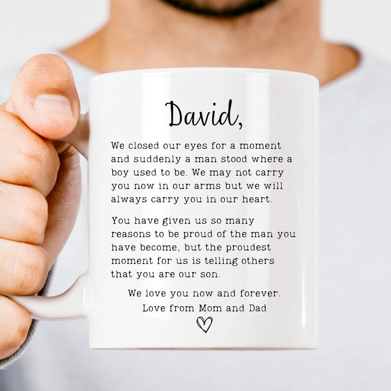K&ouml;nnte beinhalten: Eine wei&szlig;e Keramiktasse mit einer schwarz-wei&szlig;en Textnachricht, die lautet: "David, Wir schlossen unsere Augen f&uuml;r einen Moment und pl&ouml;tzlich stand ein Mann da, wo ein Junge war. Wir k&ouml;nnen dich jetzt nicht mehr in unseren Armen tragen, aber wir werden dich immer in unserem Herzen tragen. Du hast uns so viele Gr&uuml;nde gegeben, stolz auf den Mann zu sein, der du geworden bist, aber der stolzeste Moment f&uuml;r uns ist, anderen zu sagen, dass du unser Sohn bist. Wir lieben dich jetzt und f&uuml;r immer. Liebe von Mama und Papa" mit einem Herzsymbol.