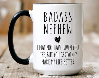 May include: White ceramic coffee mug with a black handle and black rim. The mug has black text that reads "BADASS NEPHEW" with a small heart below the text. Below the heart, the text reads "I MAY NOT HAVE GIVEN YOU LIFE, BUT YOU CERTAINLY MADE MY LIFE BETTER."