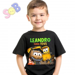 May include: Black t-shirt with the text "LEANDRO TURNS FIVE" in green and yellow. The shirt features cartoon construction vehicles. The boy is wearing the shirt and smiling.