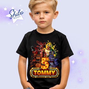 May include: Black t-shirt with a vibrant graphic of animated characters, the number 5, and the name "Tommy" in a gold-edged frame. The shirt is worn by a young boy.