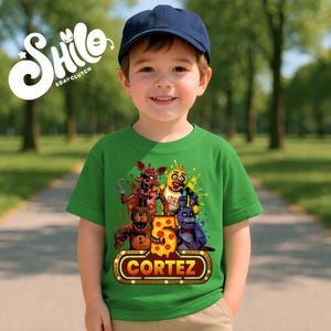May include: A green short-sleeved t-shirt featuring a graphic of four animatronic characters, a large number 5, and a marquee-style sign with the name CORTEZ. The design includes colorful paint splatters and the text SHILO BDAYCLUTCH.