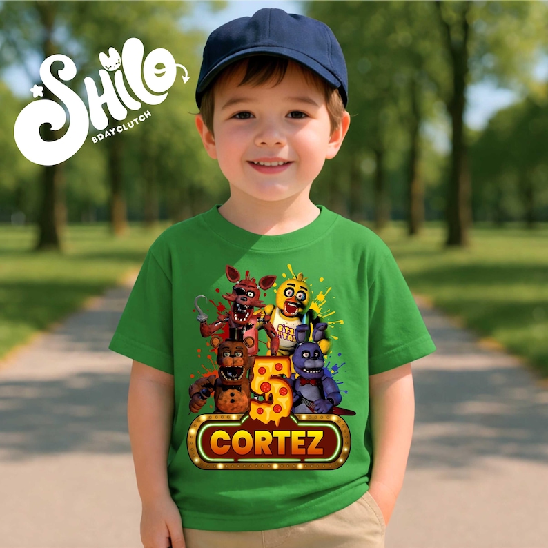 May include: A green short-sleeved t-shirt featuring a graphic of four animatronic characters, a large number 5, and a marquee-style sign with the name CORTEZ. The design includes colorful paint splatters and the text SHILO BDAYCLUTCH.