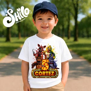 May include: A white short-sleeved t-shirt featuring a colorful graphic of four animatronic characters, a large yellow number five with pepperoni pizza patterns, and a glowing marquee sign displaying the name CORTEZ in bold orange letters.