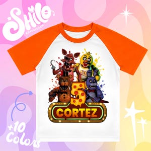 May include: A white t-shirt with orange raglan sleeves featuring a graphic of four animatronic characters, a large number five shaped like pizza, and a glowing sign with the name CORTEZ printed in bold yellow letters against a dark background.