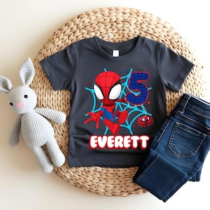 Pode incluir: Uma camiseta cinza escuro com um desenho do Homem-Aranha, o n&uacute;mero 5 e o nome "Everett" em vermelho e azul. A camisa &eacute; combinada com jeans azuis e um coelho de pel&uacute;cia cinza.