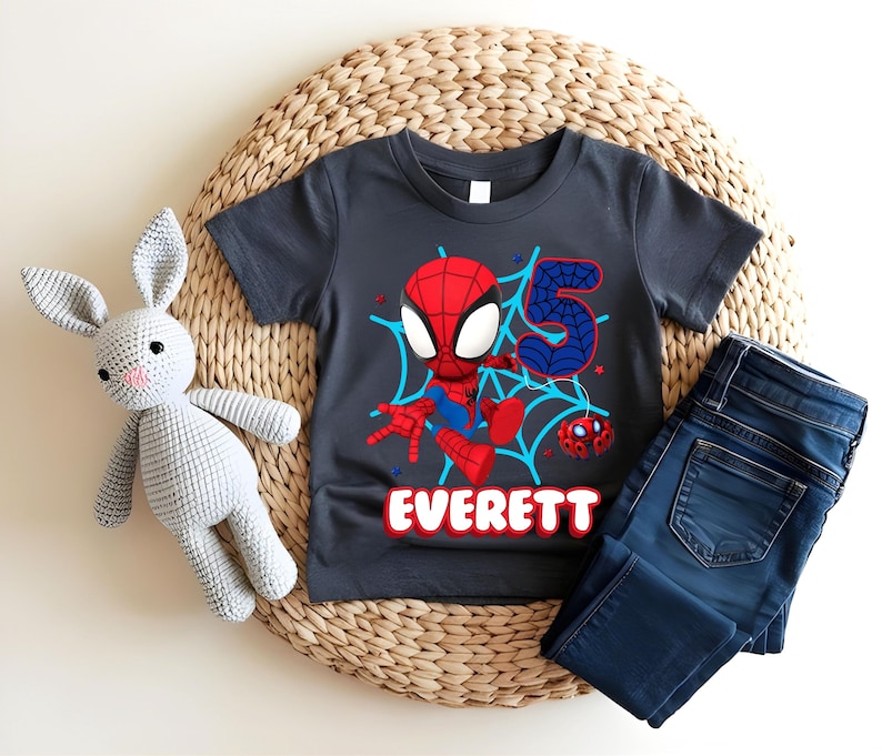 Pode incluir: Uma camiseta cinza escuro com um desenho do Homem-Aranha, o n&uacute;mero 5 e o nome "Everett" em vermelho e azul. A camisa &eacute; combinada com jeans azuis e um coelho de pel&uacute;cia cinza.
