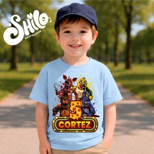 May include: A light blue t-shirt featuring a graphic print of four animated characters, a large number 5 with a pepperoni pizza pattern, and a rectangular sign with the name CORTEZ in yellow text. The shirt is worn with a dark blue baseball cap.