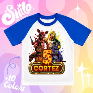 May include: A white and blue raglan t-shirt featuring a graphic of four animatronic characters from Five Nights at Freddy's, a large number 5 shaped like pizza, and a glowing marquee sign with the name CORTEZ printed in bold orange letters.