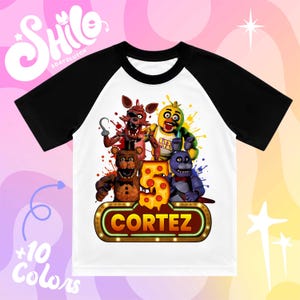 May include: A white t-shirt with black raglan sleeves featuring a graphic print of four animatronic characters, a large number five decorated with pepperoni pizza slices, and a glowing sign with the name CORTEZ in bold orange letters.