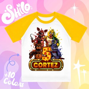 May include: A white raglan t-shirt with yellow sleeves featuring a graphic print of four animatronic characters, a large number 5 designed with pepperoni pizza slices, and a marquee-style sign with the name CORTEZ in bold yellow letters.