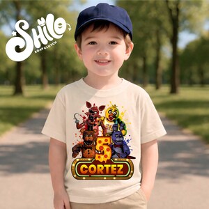 May include: A cream-colored short-sleeve t-shirt featuring a graphic print of four animatronic characters, a large number 5, and the name CORTEZ in a neon-style sign. The shirt is worn with a plain navy blue baseball cap.
