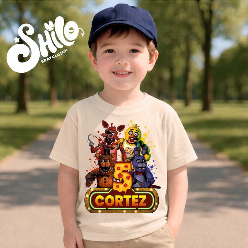 May include: A cream-colored short-sleeve t-shirt featuring a graphic print of four animatronic characters, a large number 5, and the name CORTEZ in a neon-style sign. The shirt is worn with a plain navy blue baseball cap.