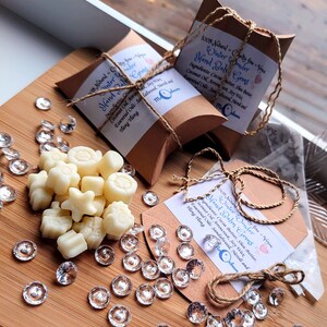 May include: A close-up of a pile of white hand balm gems in various shapes, including stars, circles, and swirls. The gems are on a wooden surface with clear glass gems scattered around. Two brown boxes with white text are visible in the background. The text on the boxes reads "100% Natural - Cruelty free - Vegan Winter Wonder Hand Balm Gems" and includes a list of ingredients.