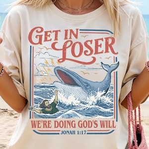 Peut inclure: T-shirt crème avec un motif d'une baleine sortant de l'océan, avec le texte "Get in Loser" au-dessus et "We're doing God's will" en dessous. Le design comprend une personne dans l'eau et le verset biblique Jonas 1:17.