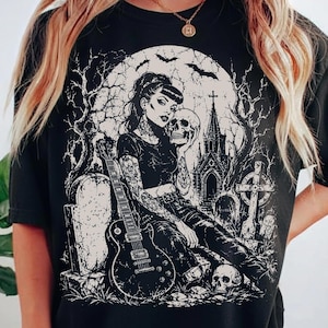 Op de afbeelding: Een zwart t-shirt met korte mouwen en een witte grafische print van een getatoeëerd persoon die op een grafsteen zit en een schedel vasthoudt, met een elektrische gitaar, een gotische kerk, kale bomen en vliegende vleermuizen op de achtergrond.