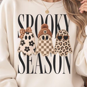 Może przedstawiać: Kremowa bluza z okrągłym dekoltem i czarnym napisem SPOOKY SEASON. Wzór zawiera trzy ilustracje duchów: jeden z brązową kokardą w kratę, drugi w brązowej czapce, a trzeci w okularach przeciwsłonecznych w kształcie serc z motywem panterki.