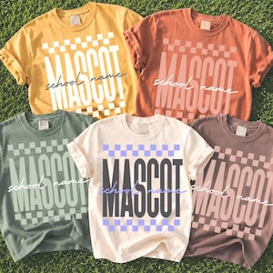 Könnte beinhalten: Fünf kurzärmelige T-Shirts in Senfgelb, Terrakotta, Salbeigrün, Creme und Mauve. Jedes T-Shirt zeigt einen karierten Rand, das Wort MASCOT in großen Blockbuchstaben und den kursiven Text school name auf der Vorderseite aufgedruckt.