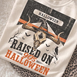 May include: An off-white t-shirt featuring a graphic of a horned animal skull wearing a witch hat. The design includes the text HALLOWEEN, RAISED ON 90's HALLOWEEN, silhouettes of bats, and a raven on a branch against a black and orange background.