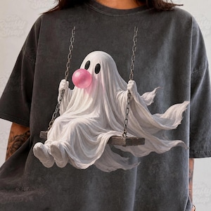 May include: A charcoal gray t-shirt featuring a graphic of a white ghost sitting on a wooden swing. The ghost has large black eyes and is blowing a round, bright pink bubble with gum. The swing is suspended by two metal chains on each side.