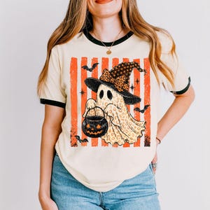 May include: A cream-coloured ringer t-shirt with black trim featuring a graphic of a ghost wearing a leopard print witch hat and holding a jack-o'-lantern bucket. The design includes orange vertical stripes, black bats, and star accents.