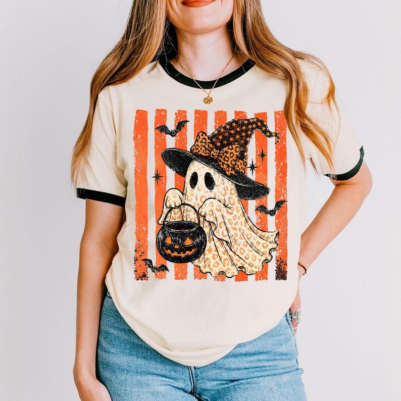 May include: A cream-coloured ringer t-shirt with black trim featuring a graphic of a ghost wearing a leopard print witch hat and holding a jack-o'-lantern bucket. The design includes orange vertical stripes, black bats, and star accents.