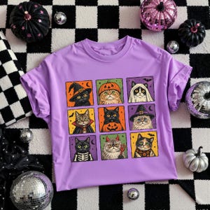K&ouml;nnte beinhalten: Ein hellviolettes T-Shirt mit einem Raster aus neun illustrierten Katzen in verschiedenen Halloween-Kost&uuml;men, darunter eine Hexe, ein K&uuml;rbis, ein Geist, ein Vampir, ein Skelett, ein Teufel sowie Katzen mit festlichen Schals und H&uuml;ten auf bunten Feldern.