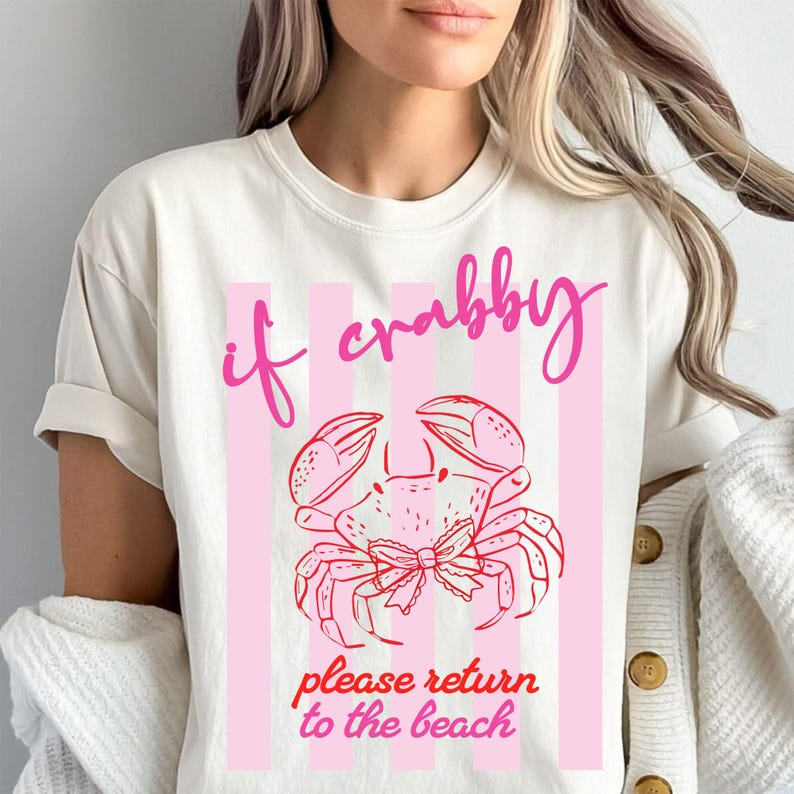 May include: Cream-colored t-shirt with a pink crab graphic and the text "if crabby please return to the beach." The crab is outlined in red and has a bow. Pink vertical stripes are behind the crab.