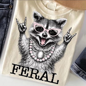 May include: A cream-coloured t-shirt with a detailed illustration of a raccoon wearing pink sunglasses and a pearl necklace, making a rock and roll hand gesture. The word "FERAL" is printed in bold, black letters below the raccoon.