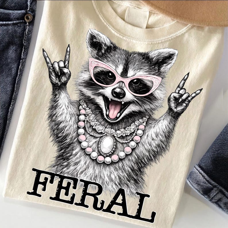 May include: A cream-coloured t-shirt with a detailed illustration of a raccoon wearing pink sunglasses and a pearl necklace, making a rock and roll hand gesture. The word "FERAL" is printed in bold, black letters below the raccoon.