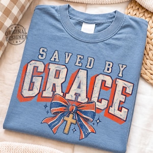 May include: A dusty blue short-sleeve t-shirt featuring the text "SAVED BY GRACE" in a distressed white font with orange drop shadows. Below the text is a graphic of a blue and orange striped bow with a gold cross charm and small star accents.