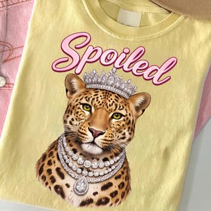 Op de afbeelding: Lichtgeel T-shirt met een luipaard met een diamanten ketting en een kroon. Het woord "Spoiled" staat in roze letters boven de luipaard. Het shirt is opgevouwen.