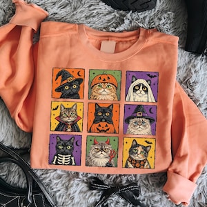 K&ouml;nnte beinhalten: Ein pfirsichfarbenes Sweatshirt mit Rundhalsausschnitt und einem Raster aus neun quadratischen Illustrationen von Katzen in verschiedenen Halloween-Kost&uuml;men, darunter eine Hexe, ein K&uuml;rbis, ein Geist, ein Vampir, ein Skelett und ein Teufel.